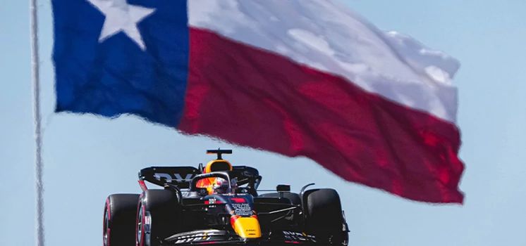 Formula 1 car racing with a large Texas state flag waving in the background.