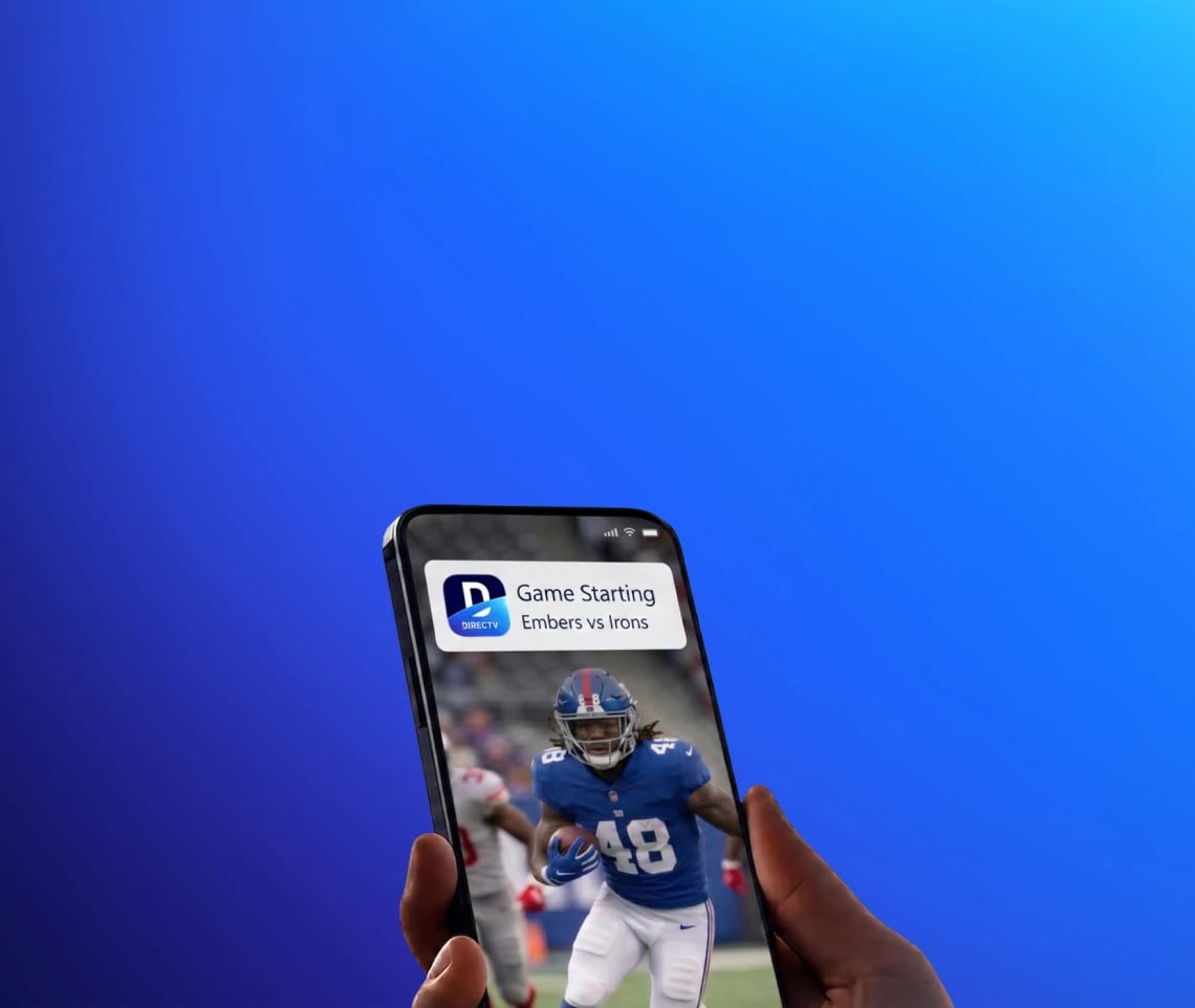 A hand holding a smartphone showing a football game-time notification alert from the DIRECTV App
