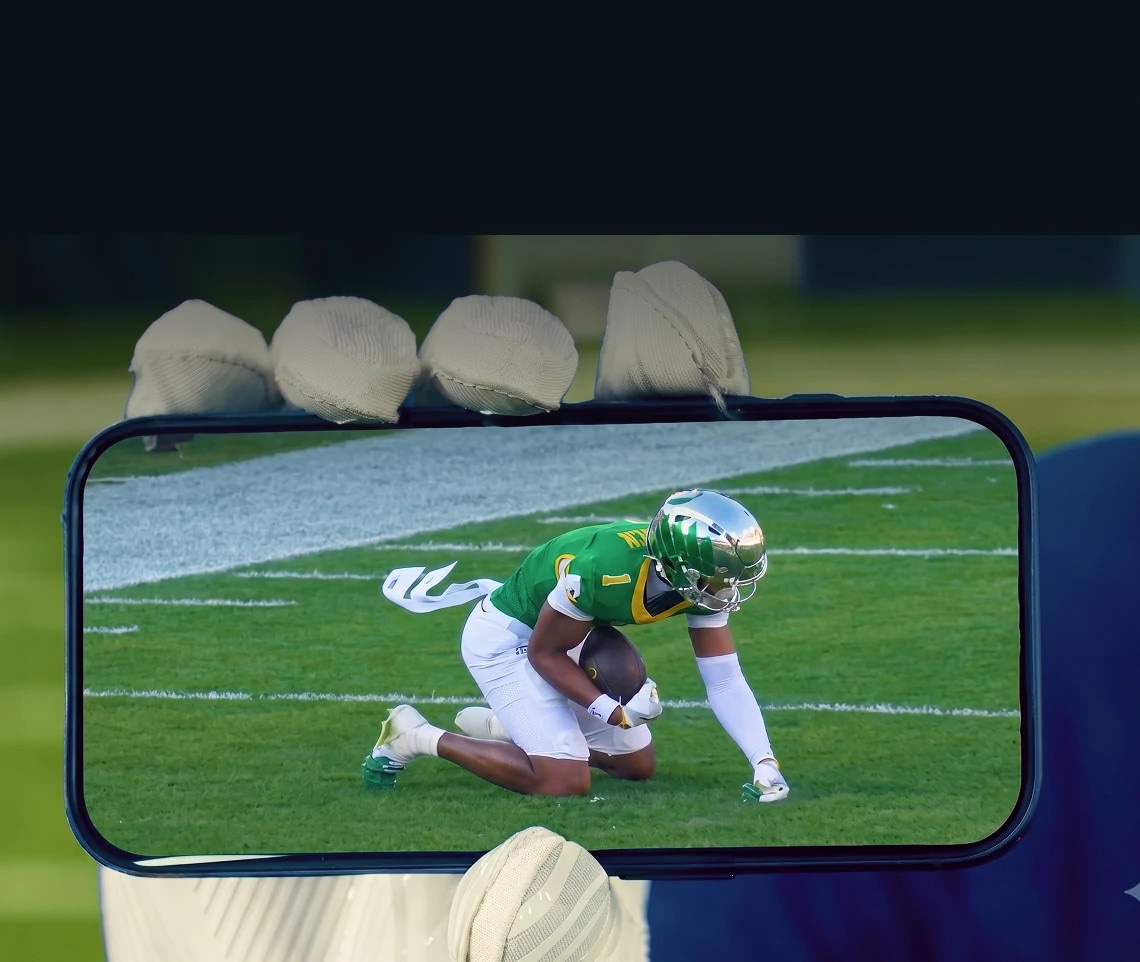 A hand holding a smartphone that shows live football on the DIRECTV app, with a football player visible on screen