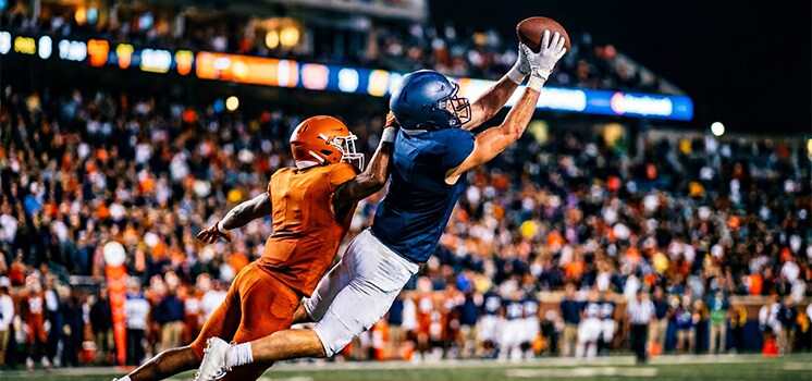 A college football player in orange and white carrying the ball past a defender in front of a packed stadium crowd.