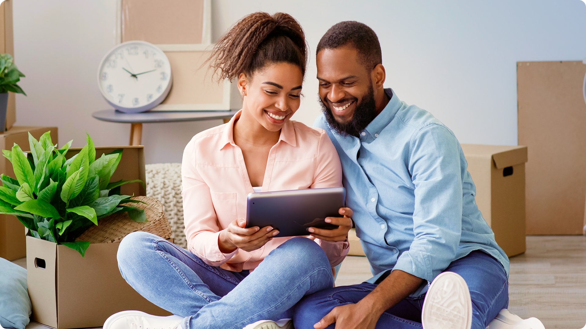 Smiling couple sitting on the floor in a new home, looking at a tablet with moving boxes nearby.