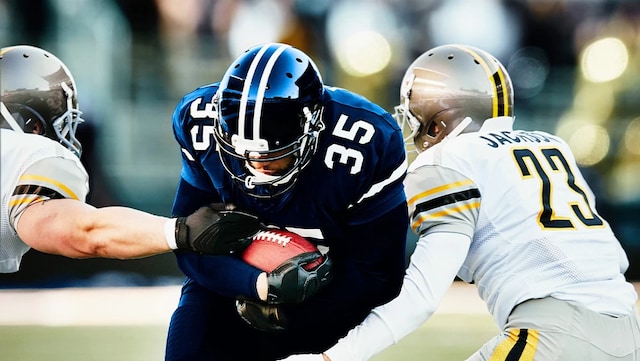 A college football player is running with a football against two opposing team players