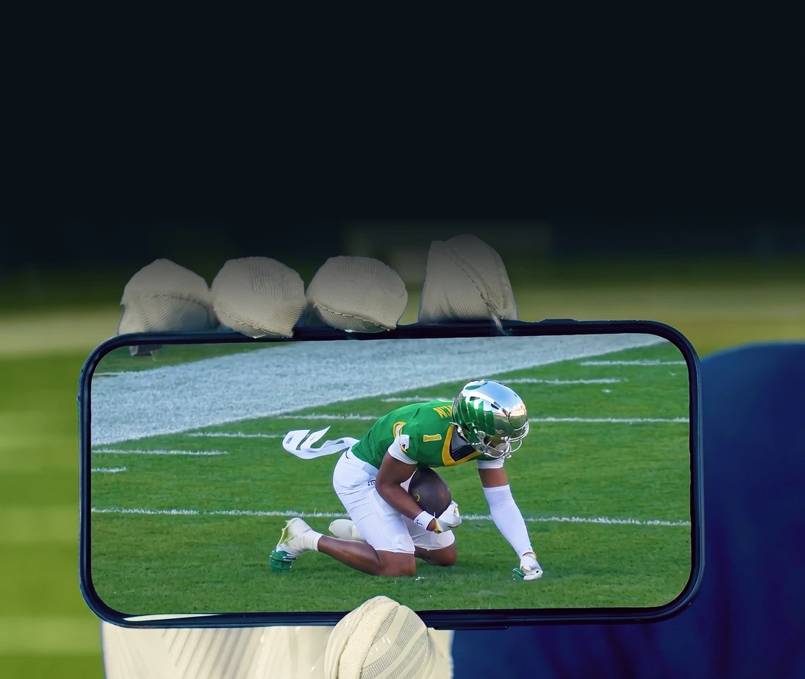 A hand holding a smartphone that shows live football on the DIRECTV app, with a football player visible on screen
