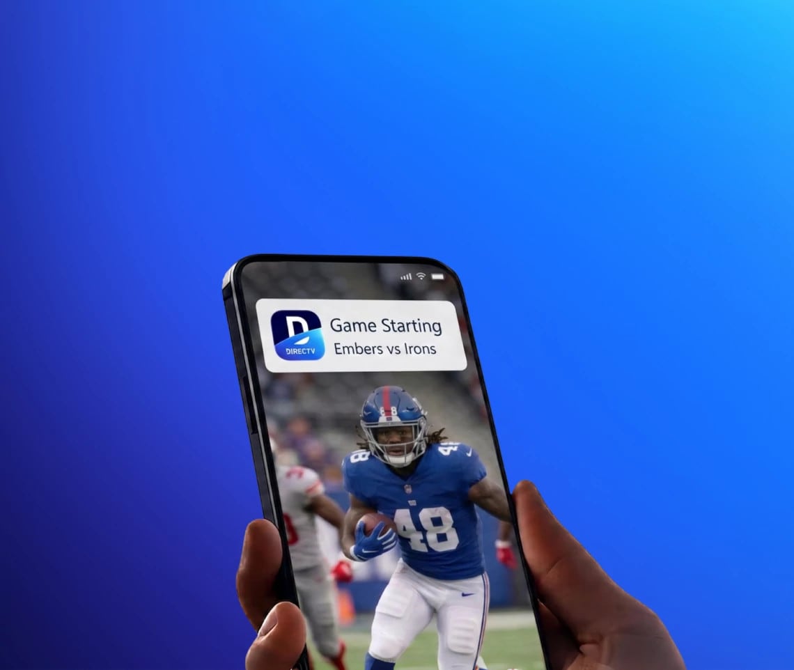 A hand holding a smartphone showing a football game-time notification alert from the DIRECTV App
